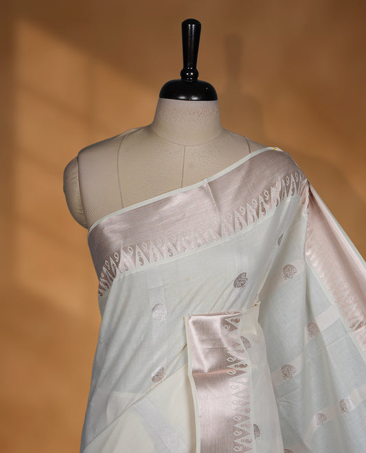 Off white colour cotton Kerala saree featuring subtle woven motifs, complemented by a soft rose gold zari border and rich traditional pallu, paired with a matching blouse.