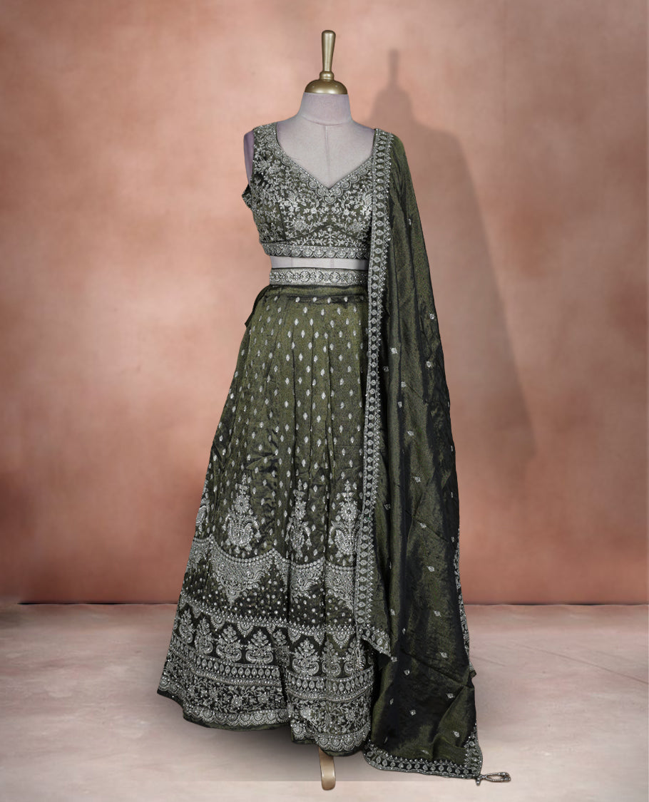 Olive green and black ombre effect lehenga set with stitched sleeveless V‑neck heavily embroidered blouse, matching stitched flared lehenga featuring intricate zari and sequin borders, and coordinated dupatta with embellished edge.