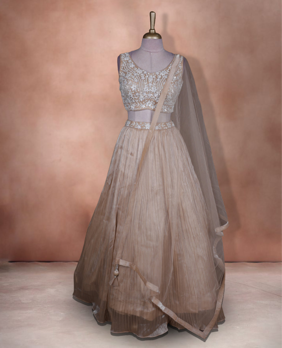 Champagne beige colour lehenga set with stitched sleeveless heavily embroidered blouse, matching stitched pleated flared lehenga, and plain sheer dupatta.