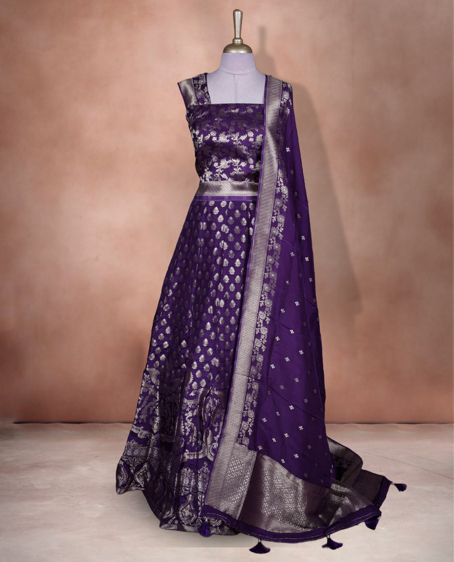 Purple colour Banarasi lehenga set with a stitched flared lehenga featuring all‑over zari butti motifs and rich woven border, paired with an unstitched floral zari blouse piece and matching dupatta with zari border and tasselled edges.