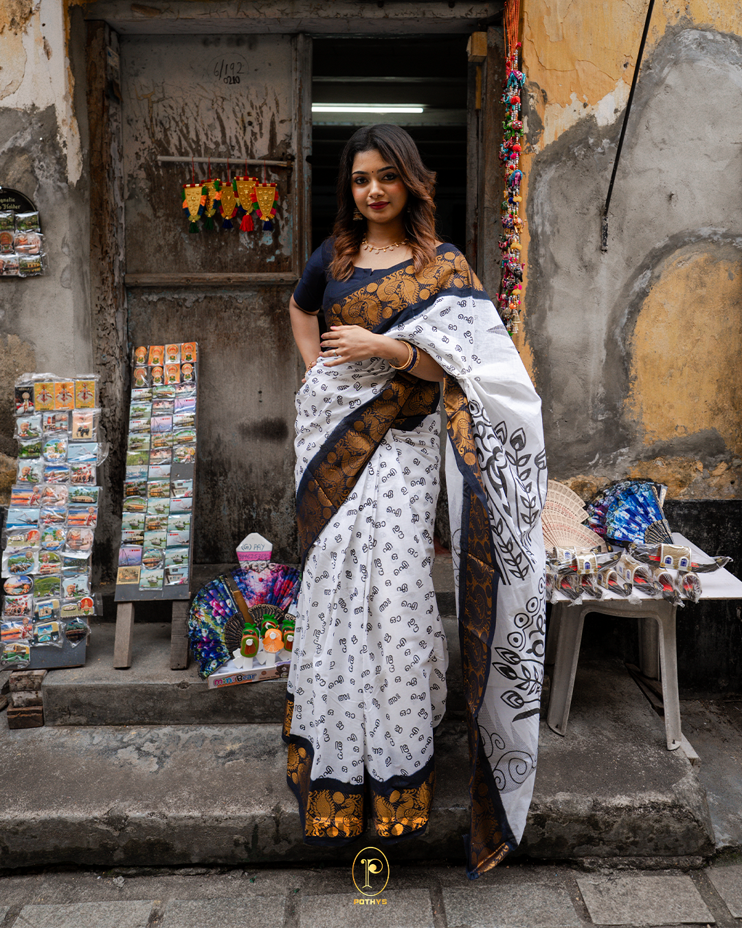 Off White Sungudi Cotton Saree featuring Malayalam alphabets on the body, a navy blue border with gold zari peacock and chakra motifs, a self-colour pallu with floral prints, and a matching blouse piece.