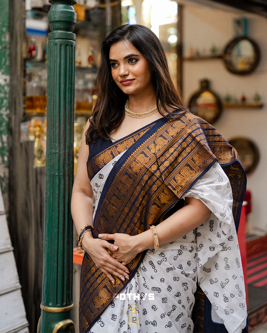 Off White Sungudi Cotton Saree featuring Malayalam alphabets on the body, a navy blue border with gold zari peacock and chakra motifs, a contrast pallu, and a matching blouse piece