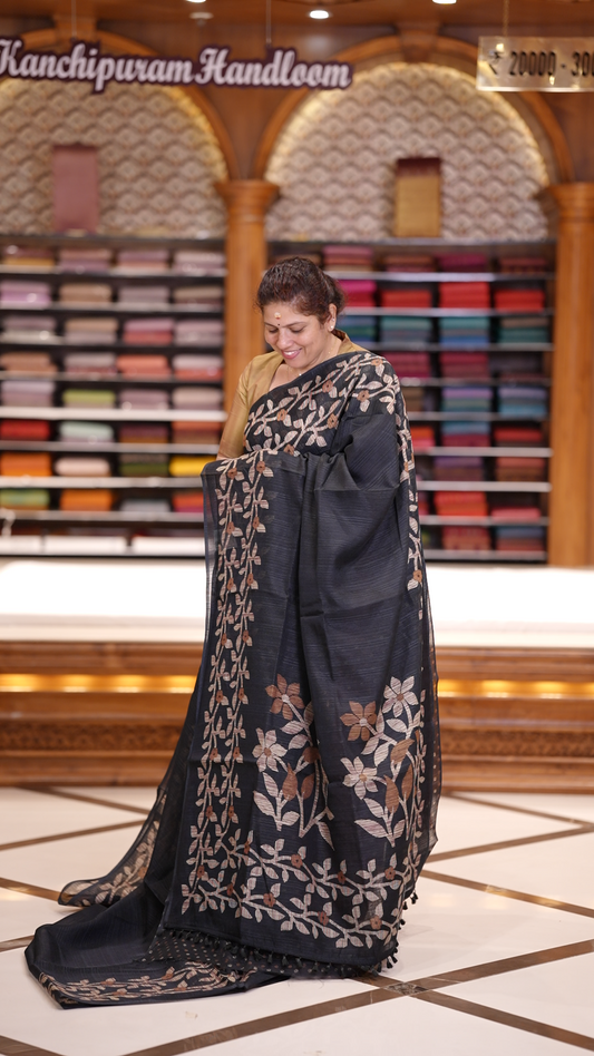 Black colour semi jute silk saree adorned with intricate floral and leaf motifs across the body & The pallu features detailed woven patterns enhancing its charm, paired beautifully with a matching floral printed blouse piece.