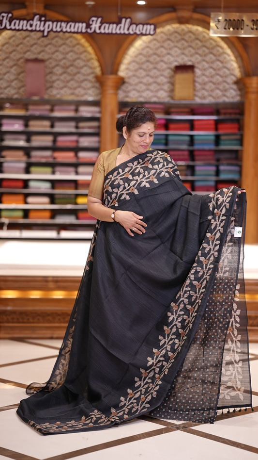 Black colour semi jute silk saree adorned with intricate floral and leaf motifs across the body & The pallu features detailed woven patterns enhancing its charm, paired beautifully with a matching floral printed blouse piece.