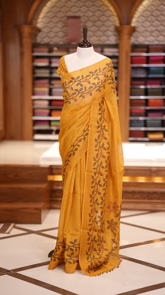 Yellow colour semi jute silk saree adorned with intricate floral and leaf motifs across the body & The pallu features detailed woven patterns enhancing its charm, paired beautifully with a matching floral printed blouse piece.