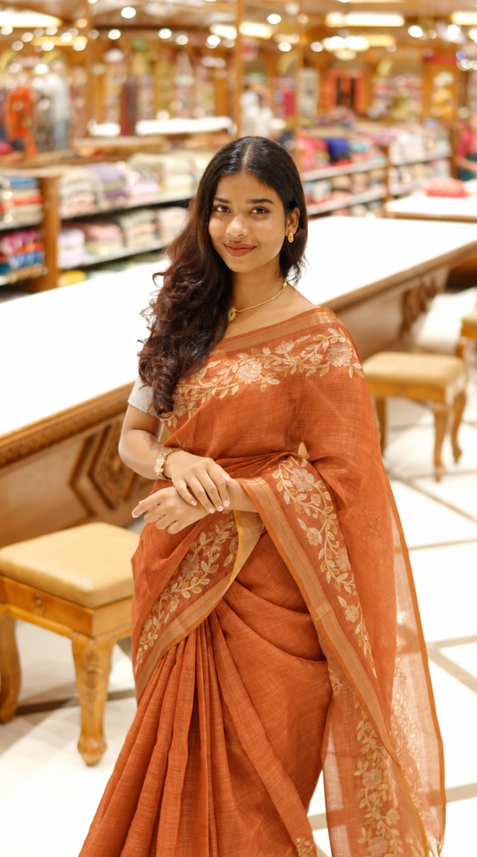 Burnt orange colour linen tissue saree featuring Floral design with Aari work & contrast simple border, comes with matching blouse piece.
