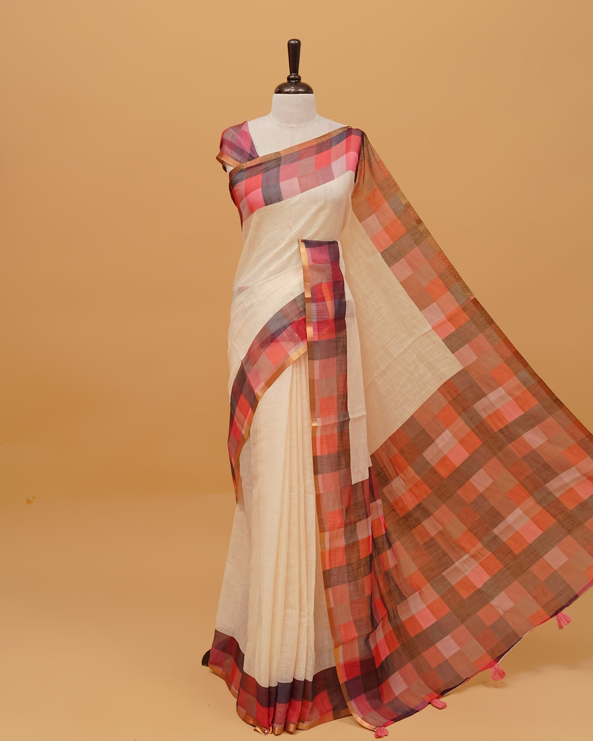 Cream colour semi linen saree featuring a plain body, a multicoloured check design with a gold tissue border, and a pallu finished with tassels. Paired with a matching blouse piece.
