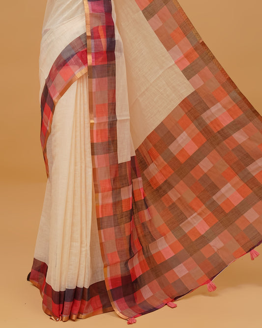 Cream colour semi linen saree featuring a plain body, a multicoloured check design with a gold tissue border, and a pallu finished with tassels. Paired with a matching blouse piece.