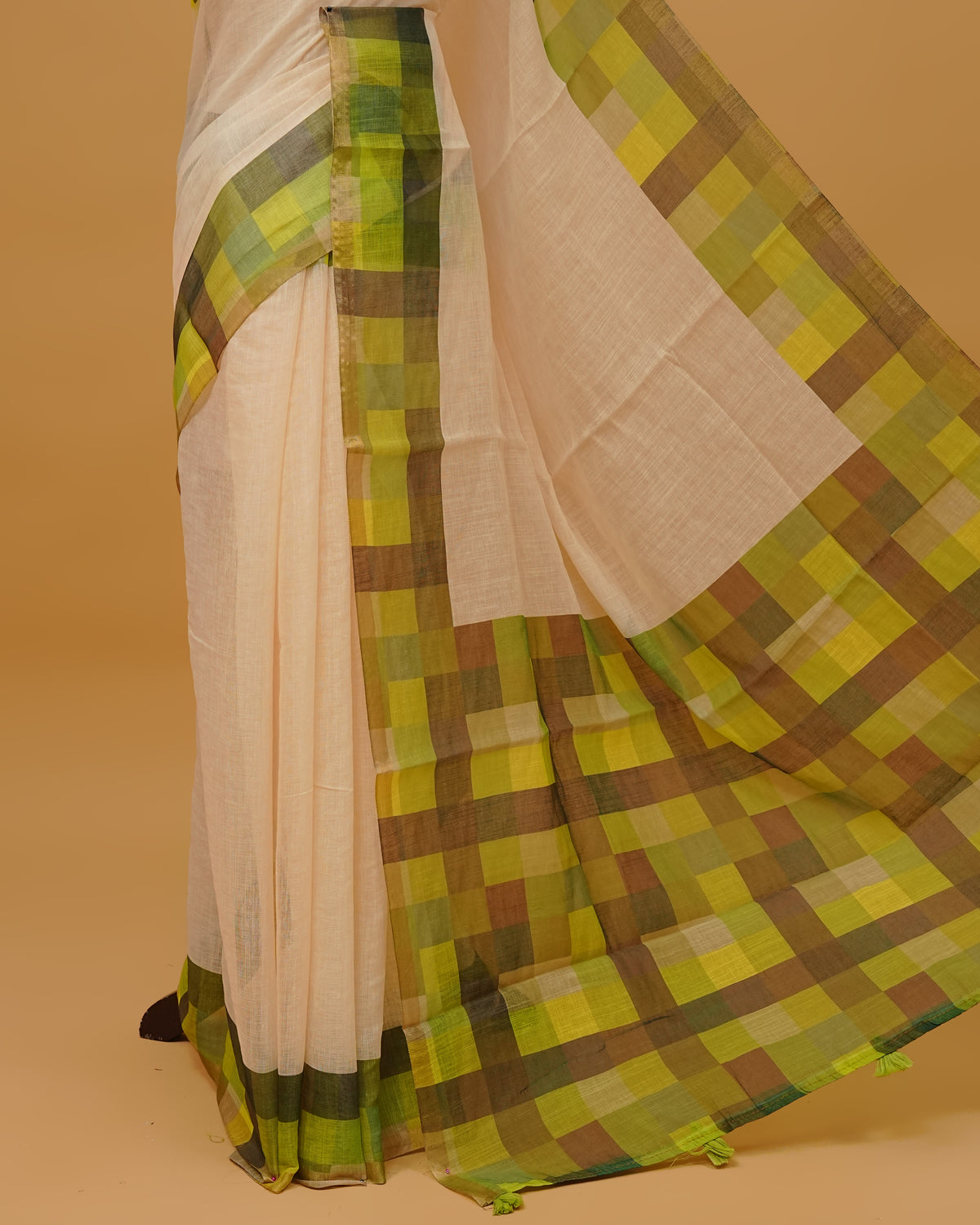 Cream colour semi linen saree featuring a plain body, a multicoloured check design with a gold tissue border, and a pallu finished with Tassels. Paired with a matching blouse piece.