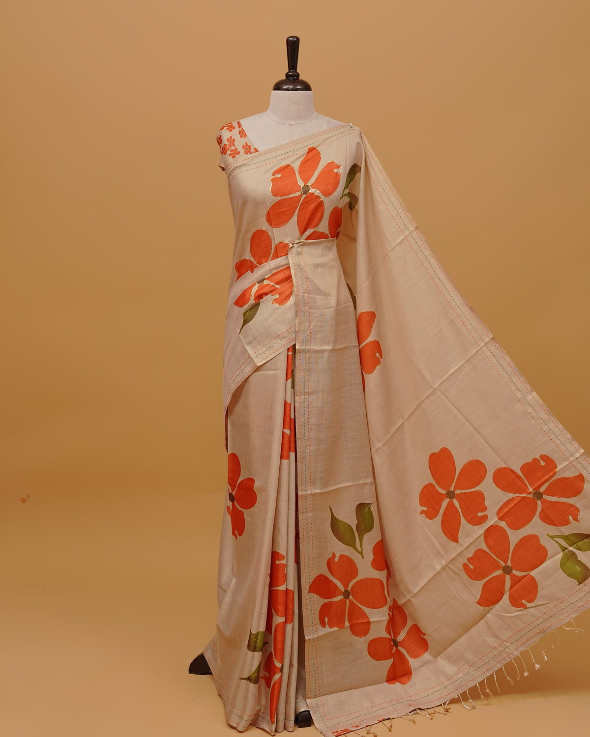 Cream colour Semi Tissue saree featuring Orange Colour Floral Motifs Design on the body, a Threaded border & a pallu finished with tassels, paired with a matching blouse piece.
