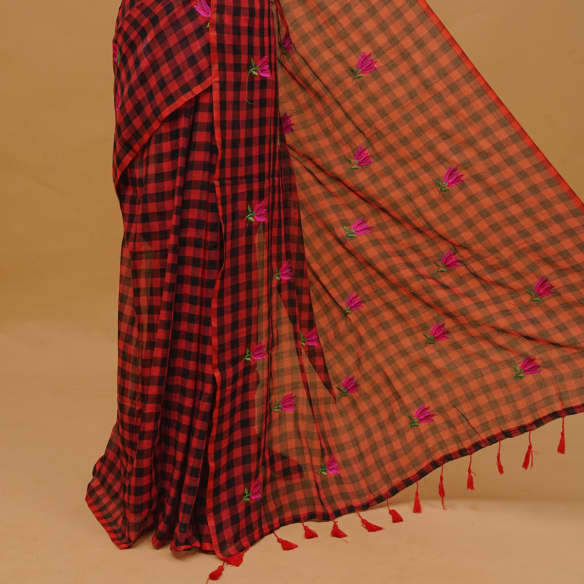 Red and black colour mul cotton saree featuring floral thread work in checked body & pallu finished with tassels. Paired with a matching blouse piece.