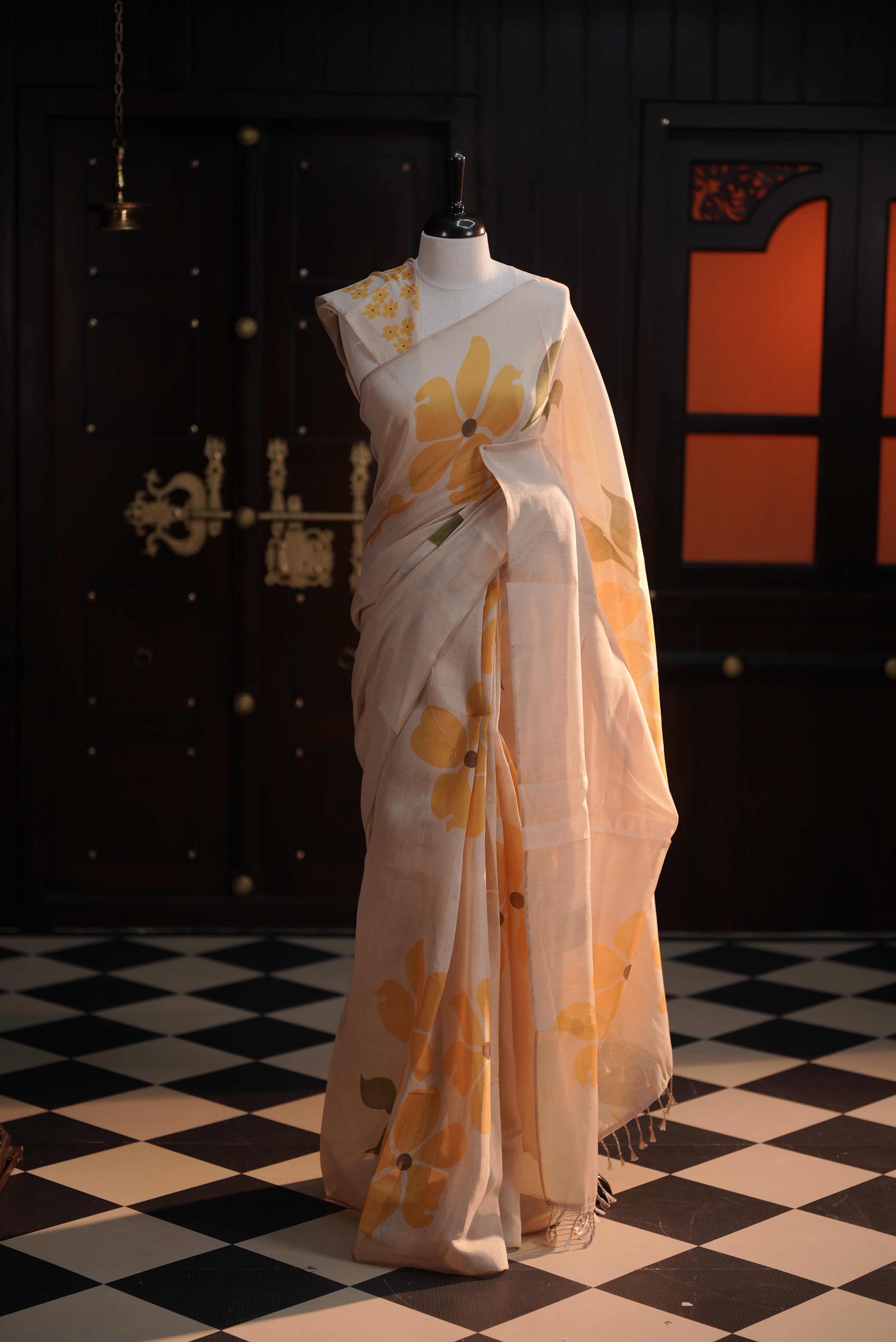 Beige colour semi soft tissue saree adorned with bold Yellow floral prints across the body, designed without a border, paired with a matching blouse piece featuring delicate flower motifs.