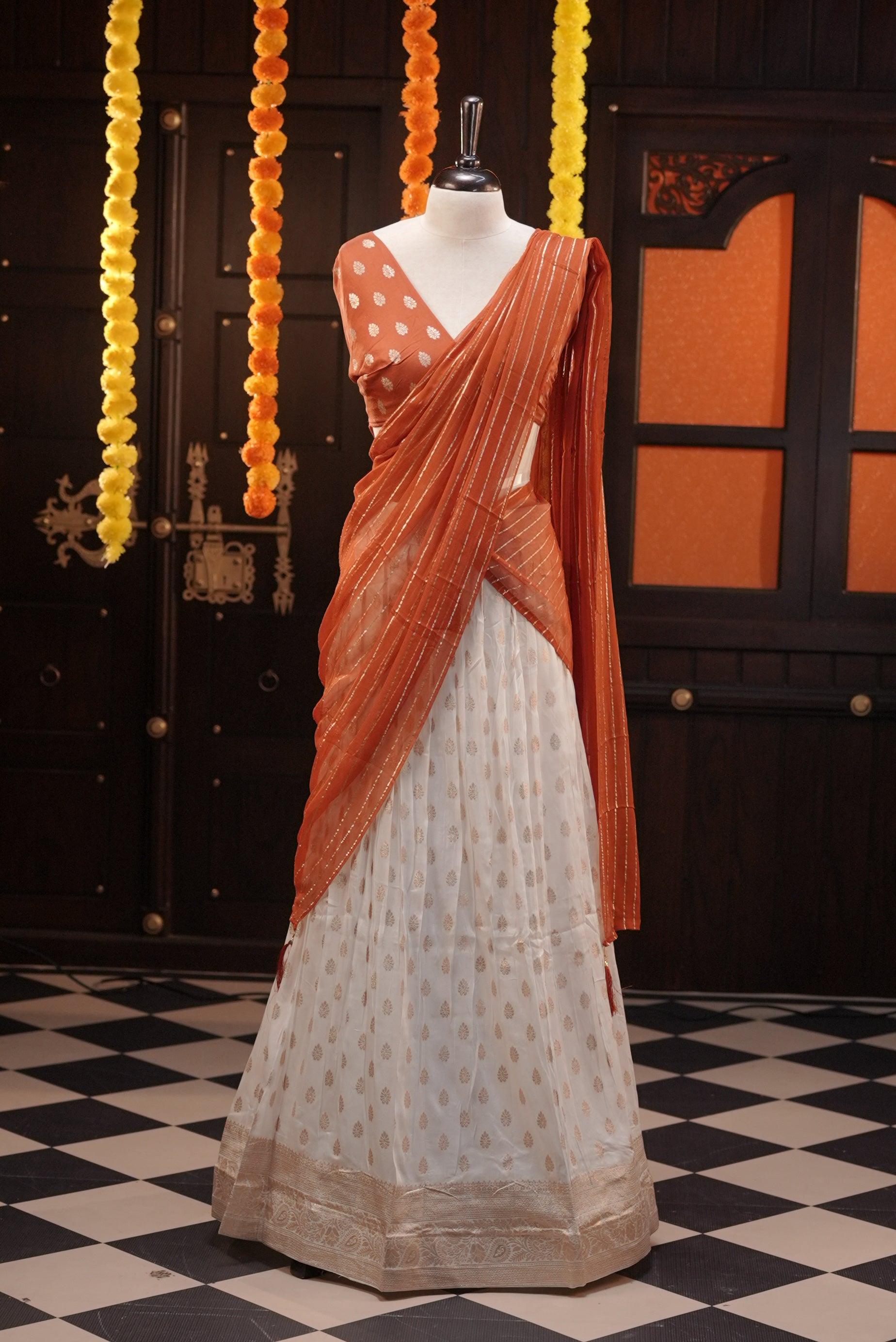 Off white colour Semi Stitched Banaras Dhavani Set Featuring off white Skirt with Gold zari buttas & Traditional border, paired with Orange colour Blouse material with gold zari buttas, finishing with same colour with gold zari Stripped Dupatta.