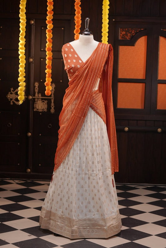Off white colour Semi Stitched Banaras Dhavani Set Featuring off white Skirt with Gold zari buttas & Traditional border, paired with Orange colour Blouse material with gold zari buttas, finishing with same colour with gold zari Stripped Dupatta.