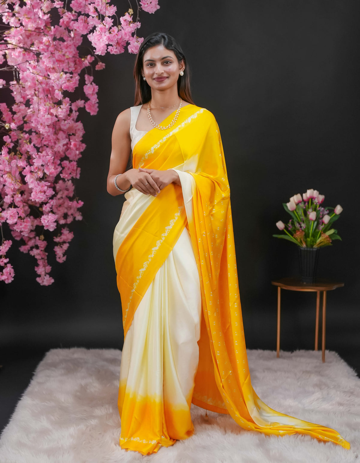 Yellow coloured modal satin saree featuring a plain cream coloured body, complemented by a double shaded border and a Bandhani designed pallu with jhumka tassels. Paired with a matching contrast coloured blouse piece.