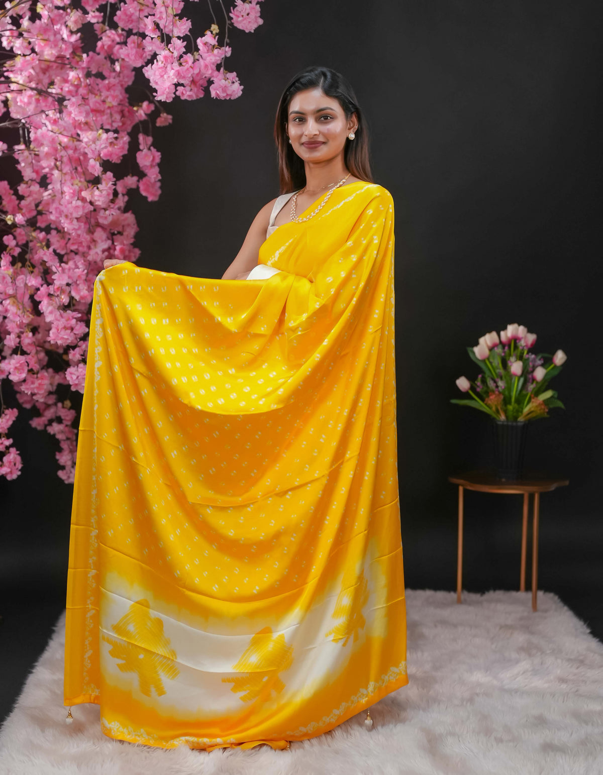 Yellow coloured modal satin saree featuring a plain cream coloured body, complemented by a double shaded border and a Bandhani designed pallu with jhumka tassels. Paired with a matching contrast coloured blouse piece.
