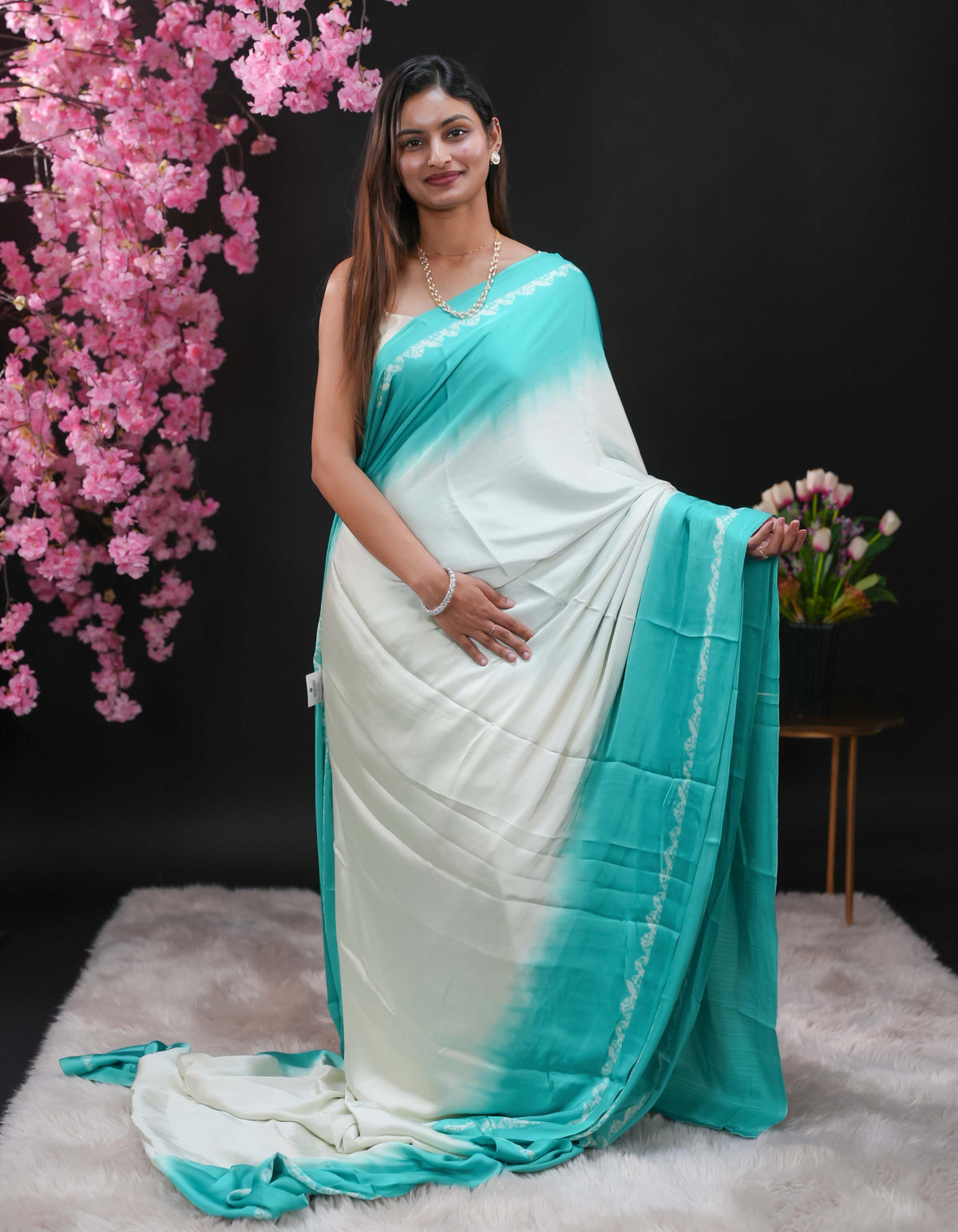 Teal coloured modal satin saree featuring a plain cream coloured body, complemented by a double shaded border and a Bandhani designed pallu with jhumka tassels. Paired with a matching contrast coloured blouse piece.