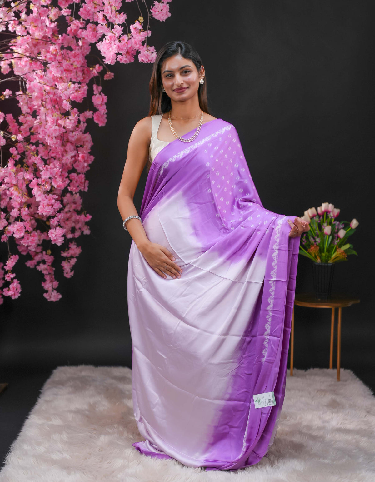 Lavender coloured modal satin saree featuring a plain cream coloured body, complemented by a double shaded border and a Bandhani designed pallu with jhumka tassels. Paired with a matching contrast coloured blouse piece.