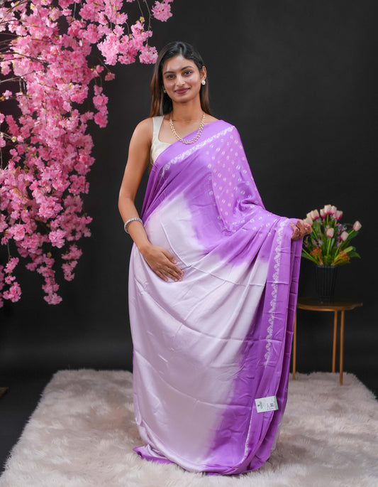 Lavender coloured modal satin saree featuring a plain cream coloured body, complemented by a double shaded border and a Bandhani designed pallu with jhumka tassels. Paired with a matching contrast coloured blouse piece.