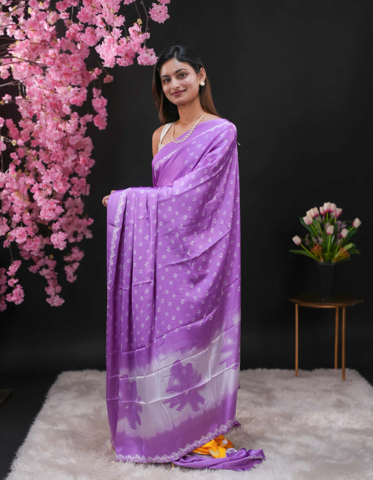 Lavender coloured modal satin saree featuring a plain cream coloured body, complemented by a double shaded border and a Bandhani designed pallu with jhumka tassels. Paired with a matching contrast coloured blouse piece.