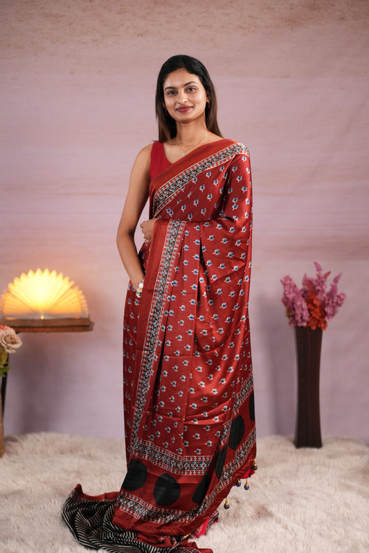 Coffee Brown and Black colour satin modal saree featuring stripes on the body, Ajrakh prints on the border & polka dots with jhumka tassels on the pallu, paired with a matching Ajrakh printed blouse piece.