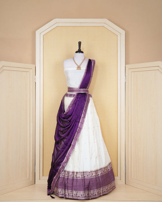 Cream coloured semi silk semi stitched dhavani set featuring gold zari buttas on the body with a contrast purple skirt with a gold zari elephant border, matching blouse piece and dupatta