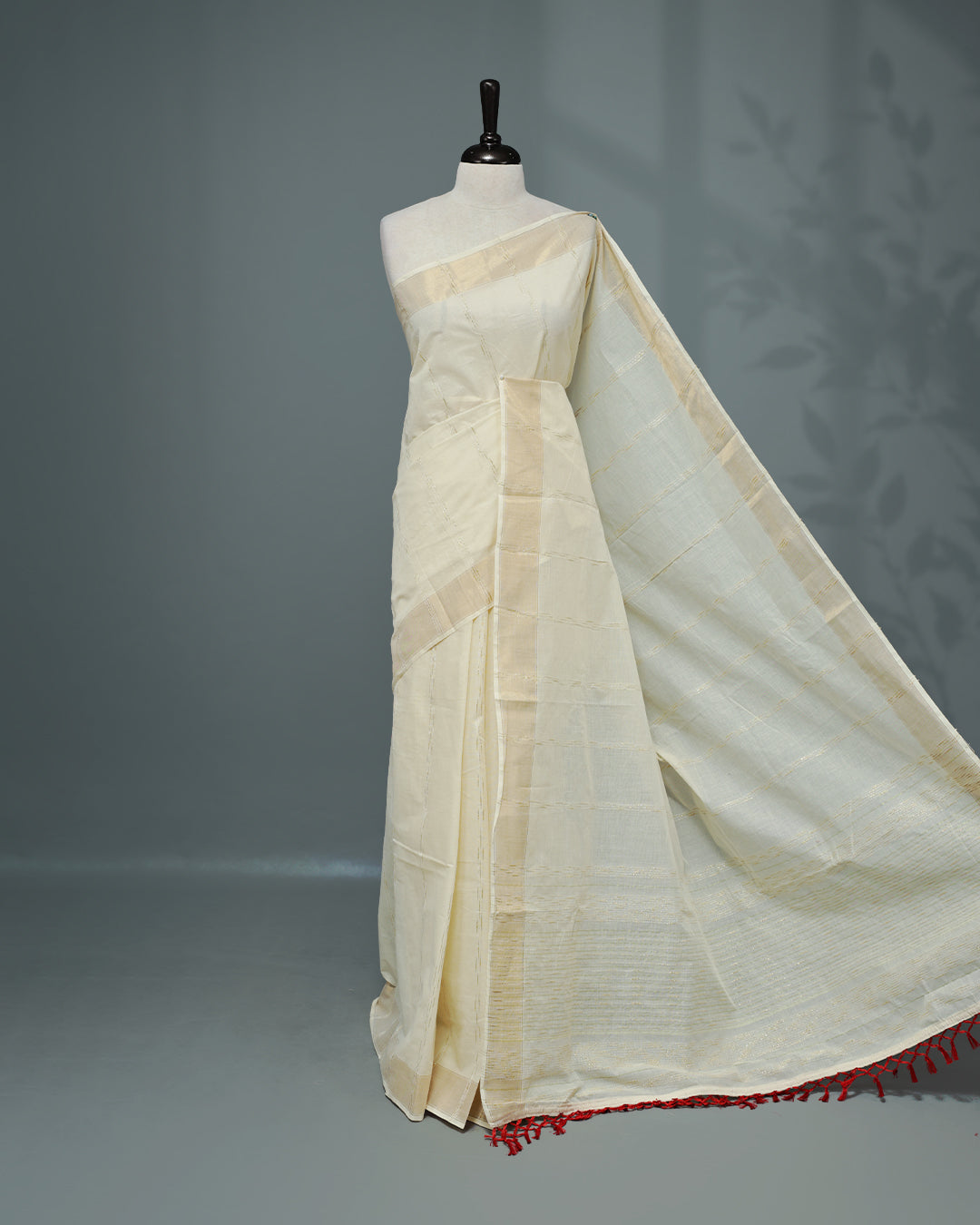 Cream colour Cotton kerala saree featuring Gold Embroidery strips on the body, complemented by a gold zari Border, red elegant tassels, paired with a matching blouse piece.