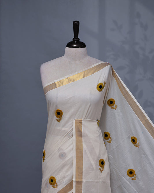 Cream colour Cotton kerala saree featuring Sunflower Embroidery work on the body, complemented by a gold zari Border, paired with a matching blouse piece.