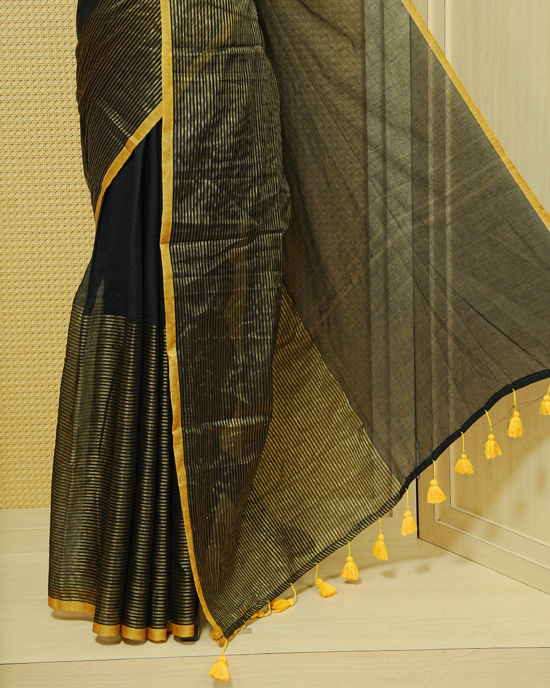 Black Mul Cotton Saree featuring an elegant plain body, beautifully complemented by a gold zari striped border and mustard-yellow tassels.