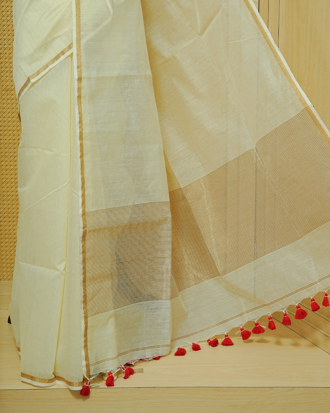 Cream Mul Cotton Saree featuring a graceful plain body with a subtle gold zari border and a finely woven striped pallu. Finished with vibrant red tassels