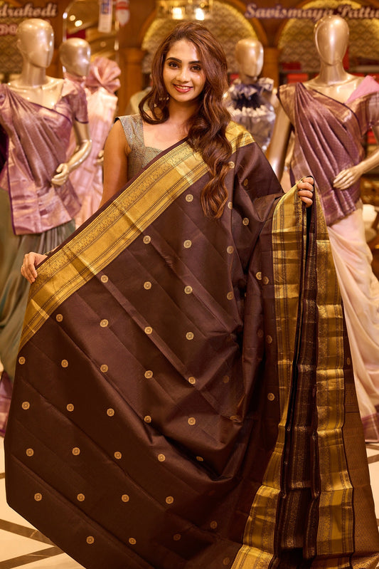 Graceful deep chocolate brown colour Kancheepuram silk saree adorned with delicate gold zari motifs across the body, beautifully finished with a broad traditional gold zari border, exuding timeless elegance and paired with a matching blouse piece