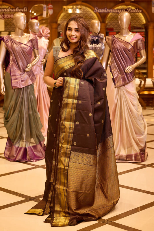 Graceful deep chocolate brown colour Kancheepuram silk saree adorned with delicate gold zari motifs across the body, beautifully finished with a broad traditional gold zari border, exuding timeless elegance and paired with a matching blouse piece
