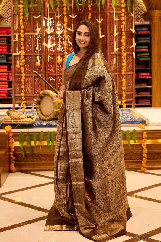 Dark chocolate colour Kancheepuram silk saree featuring intricate gold zari geometric and floral weaving throughout, completed with a rich gold zari border and grand pallu, paired with a matching blouse