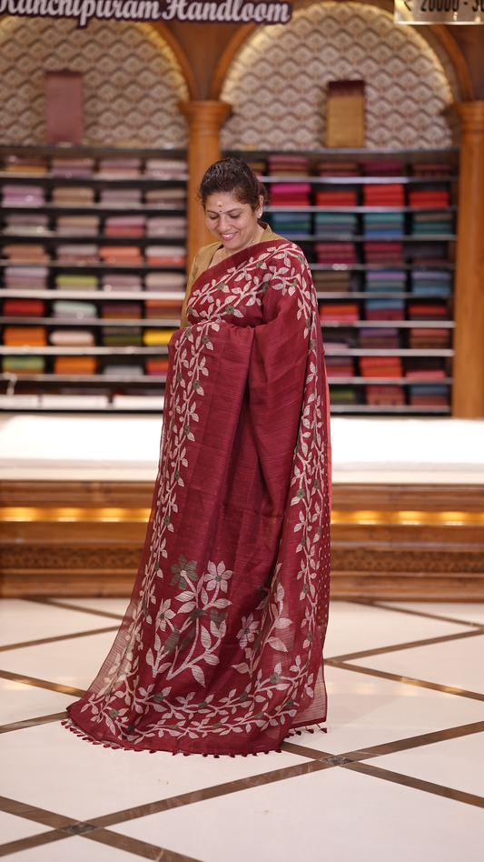 Maroon colour semi jute silk saree adorned with intricate floral and leaf motifs across the body & The pallu features detailed woven patterns enhancing its charm, paired beautifully with a matching floral printed blouse piece.