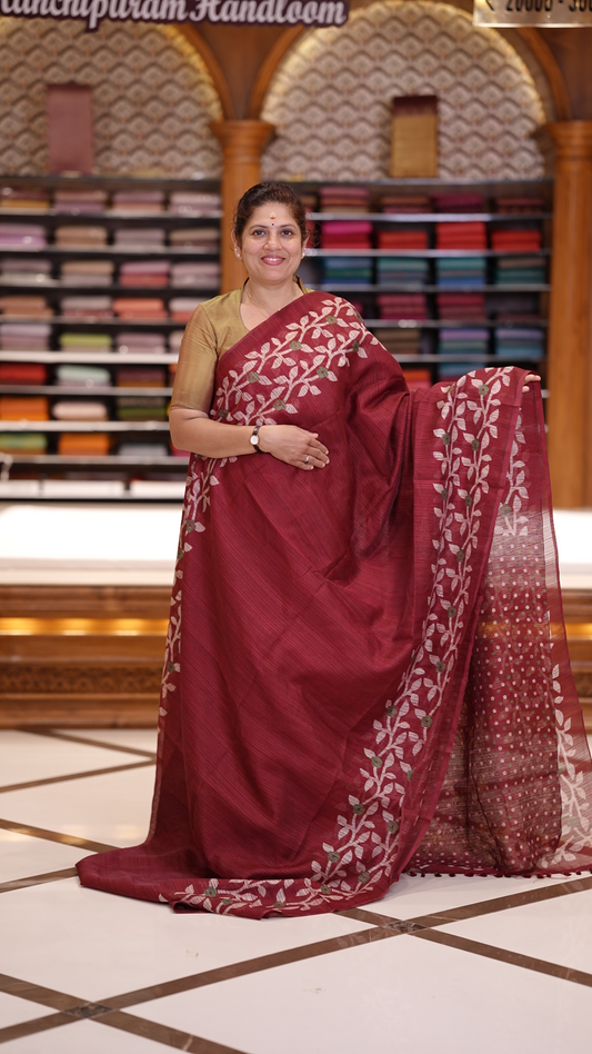 Maroon colour semi jute silk saree adorned with intricate floral and leaf motifs across the body & The pallu features detailed woven patterns enhancing its charm, paired beautifully with a matching floral printed blouse piece.