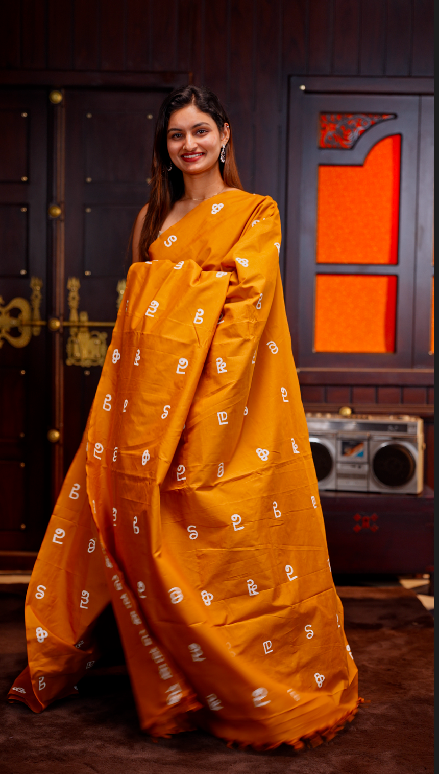 Mustard Yellow colour Poly cotton saree featuring Malayalam Letters Print on body, pallu with tassels, Contrast Colour Blouse piece.