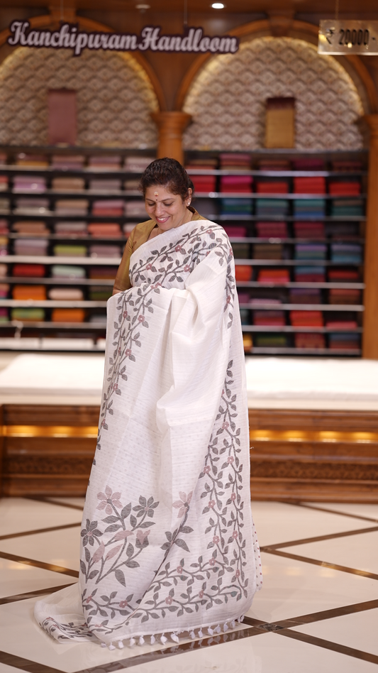 White colour semi jute silk saree adorned with intricate floral and leaf motifs across the body & The pallu features detailed woven patterns enhancing its charm, paired beautifully with a matching floral printed blouse piece.