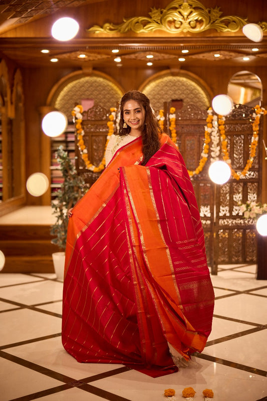 Pure silk saree featuring a rich red body with golden zari stripes, contrasted by an orange pallu and finely woven zari border.