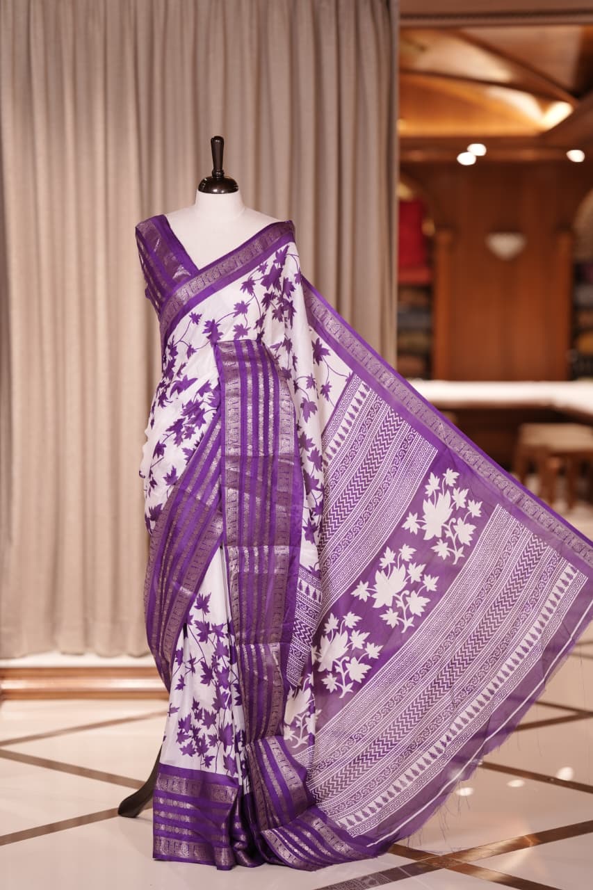 White colour semi tussar silk saree adorned with lavender floral prints, highlighted by lavender Colour traditional border Paired with a matching blouse and a beautifully printed pallu for classic elegance.