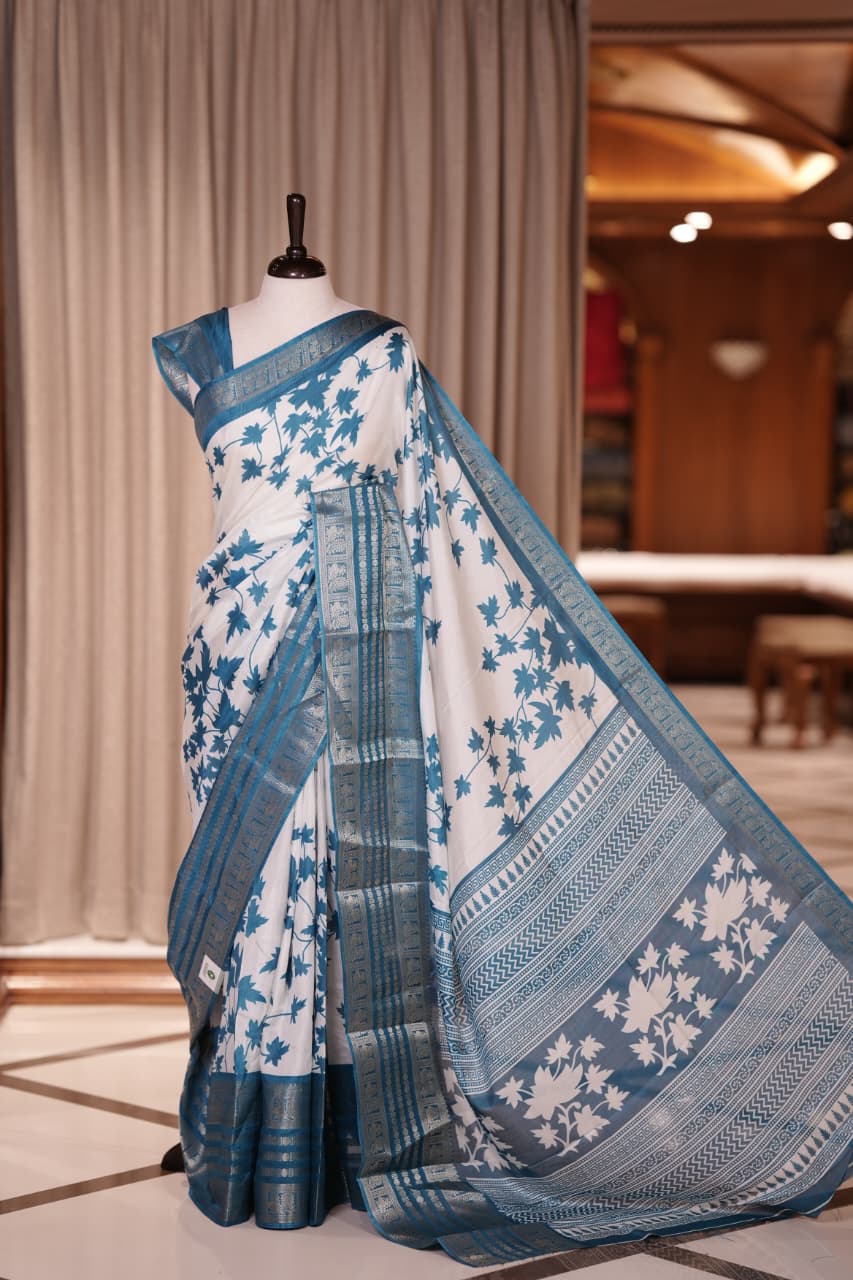 White colour semi tussar silk saree adorned with Blue floral prints, highlighted by Blue Colour traditional border Paired with a matching blouse and a beautifully printed pallu for classic elegance.