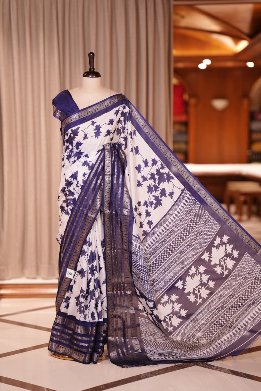 White colour semi tussar silk saree adorned with purple floral prints, highlighted by purple Colour traditional border Paired with a matching blouse and a beautifully printed pallu for classic elegance.