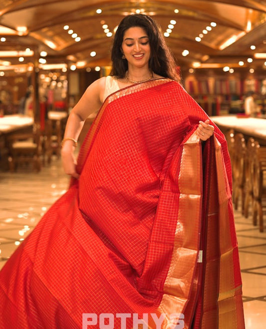 Red colour semi banaras lichi silk saree featuring checked patterned body, complemented by gold zari border, paired with matching blouse piece.