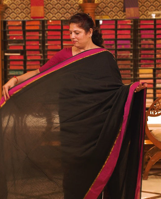 Black colour Mul Cotton saree featuring a simple plain body with a contrast border, offering a classic and elegant look. Comes without a blouse piece.