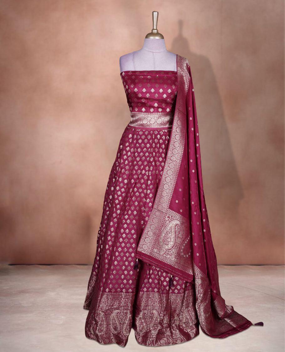 Wine colour Banarasi lehenga set with a stitched flared lehenga featuring all‑over zari butti motifs and rich woven border, paired with an unstitched floral zari blouse piece and matching dupatta with zari border and tasselled edges.