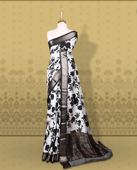 White colour saree adorned with bold black floral prints and a rich black zari border and stripped checkered pallu & Comes with a matching blouse piece.