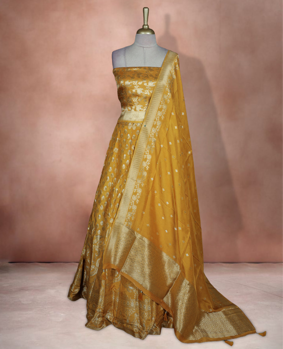 Yellow colour Banarasi lehenga set with a stitched flared lehenga featuring all‑over zari butti motifs and rich woven border, paired with an unstitched floral zari blouse piece and matching dupatta with zari border and tasselled edges.